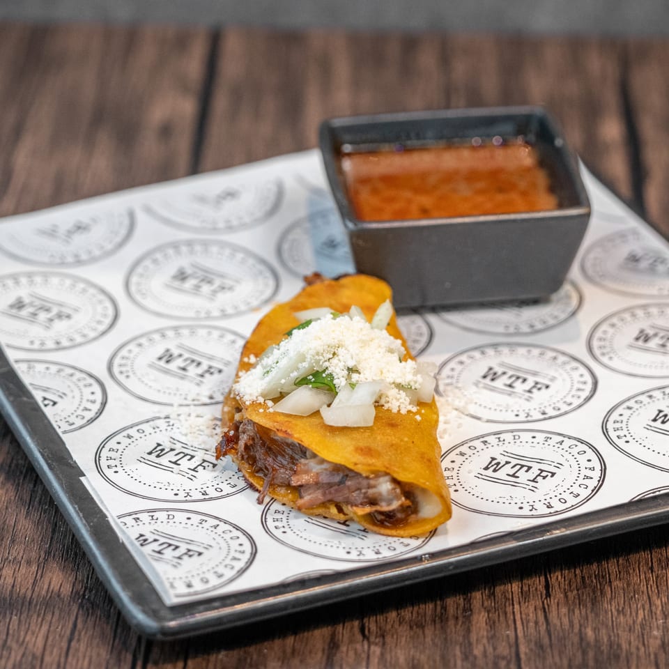 A World Taco Factory birria taco with cheese and onions, served with a side of consommé.