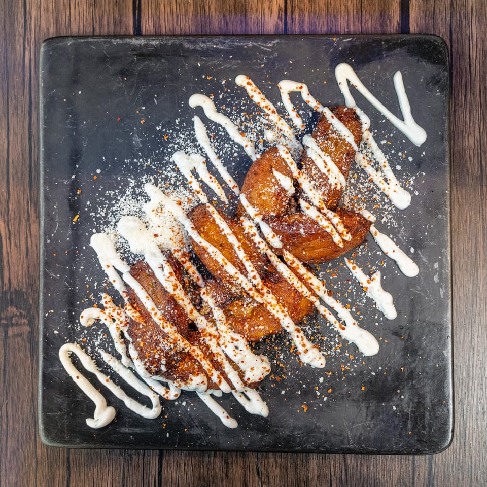 Fried plantains drizzled with white sauce and sprinkled with spices on a dark plate.