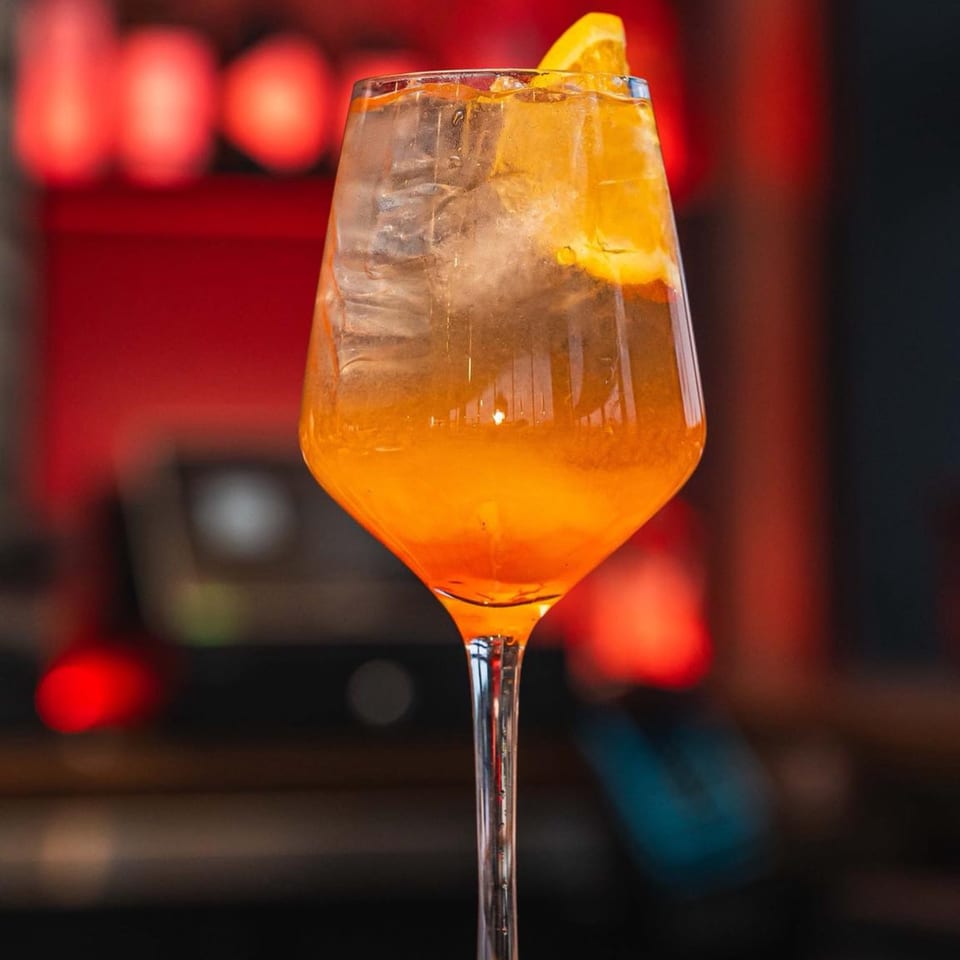 A vibrant orange cocktail with ice and an orange slice, against a blurred red background.