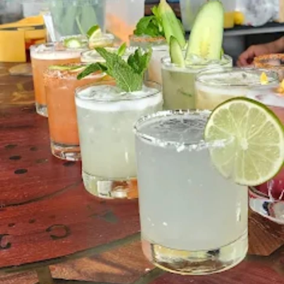 A row of World Taco Factory margaritas, some garnished with lime, mint, or cucumber, on a wooden bar.
