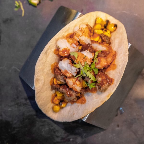 A World Taco Factory taco with fish, steak, corn, sauce, and cilantro on a dark surface.