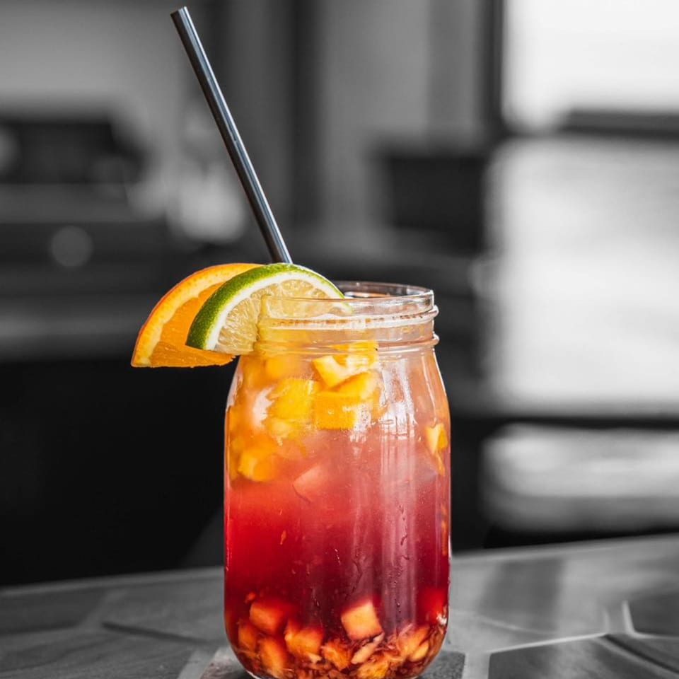 A vibrant red and orange fruit drink in a mason jar with a straw, garnished with citrus slices.