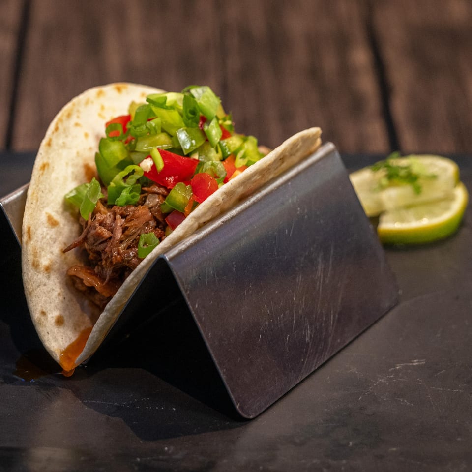 A World Taco Factory taco with shredded meat, green and red peppers, and lime wedges.