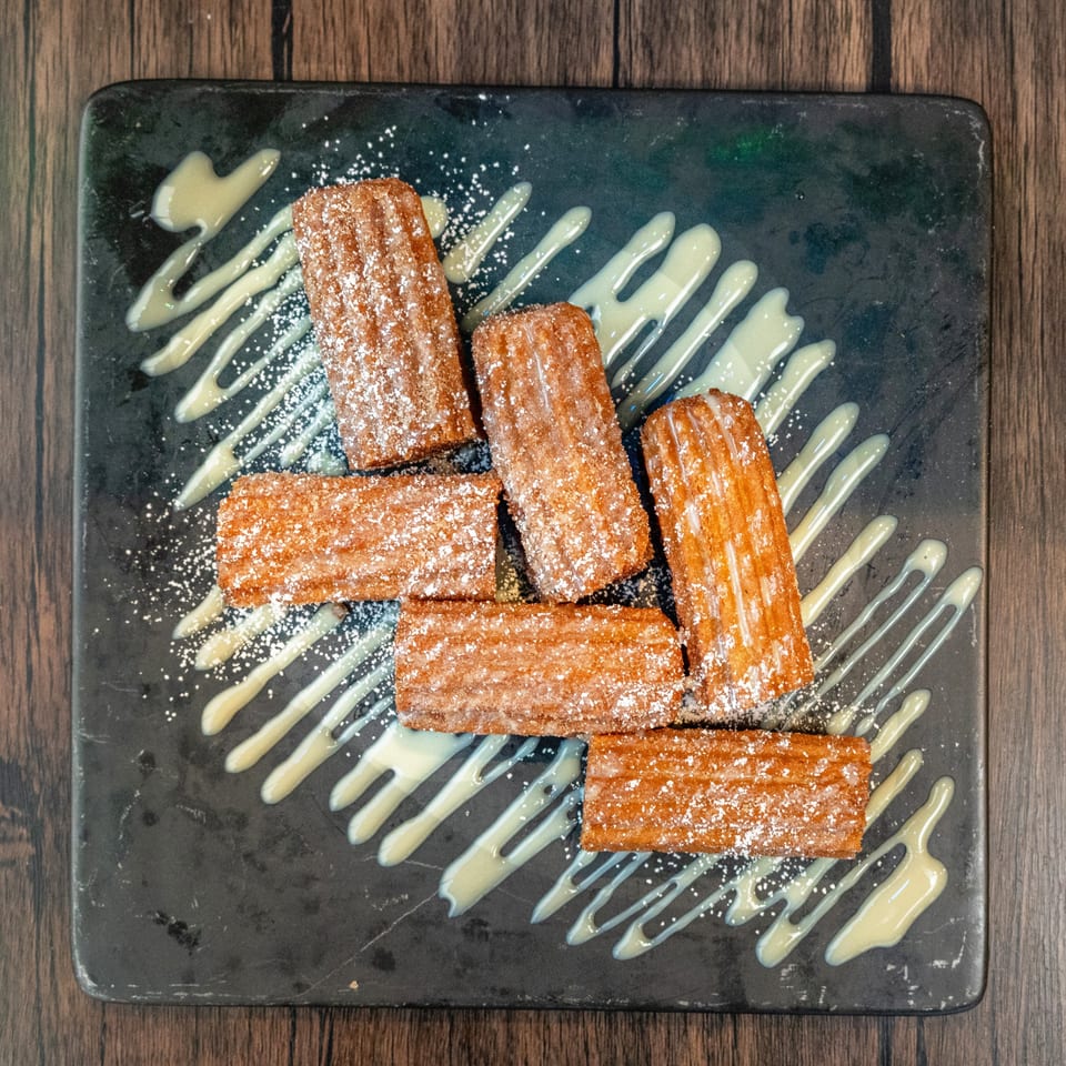 Six churros dusted with powdered sugar and drizzled with white icing on a dark plate.