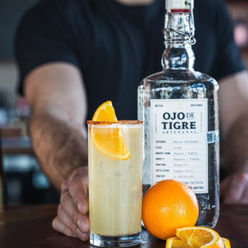 A bartender's hand next to a mezcal cocktail garnished with an orange slice and a bottle of Ojo de Tigre.