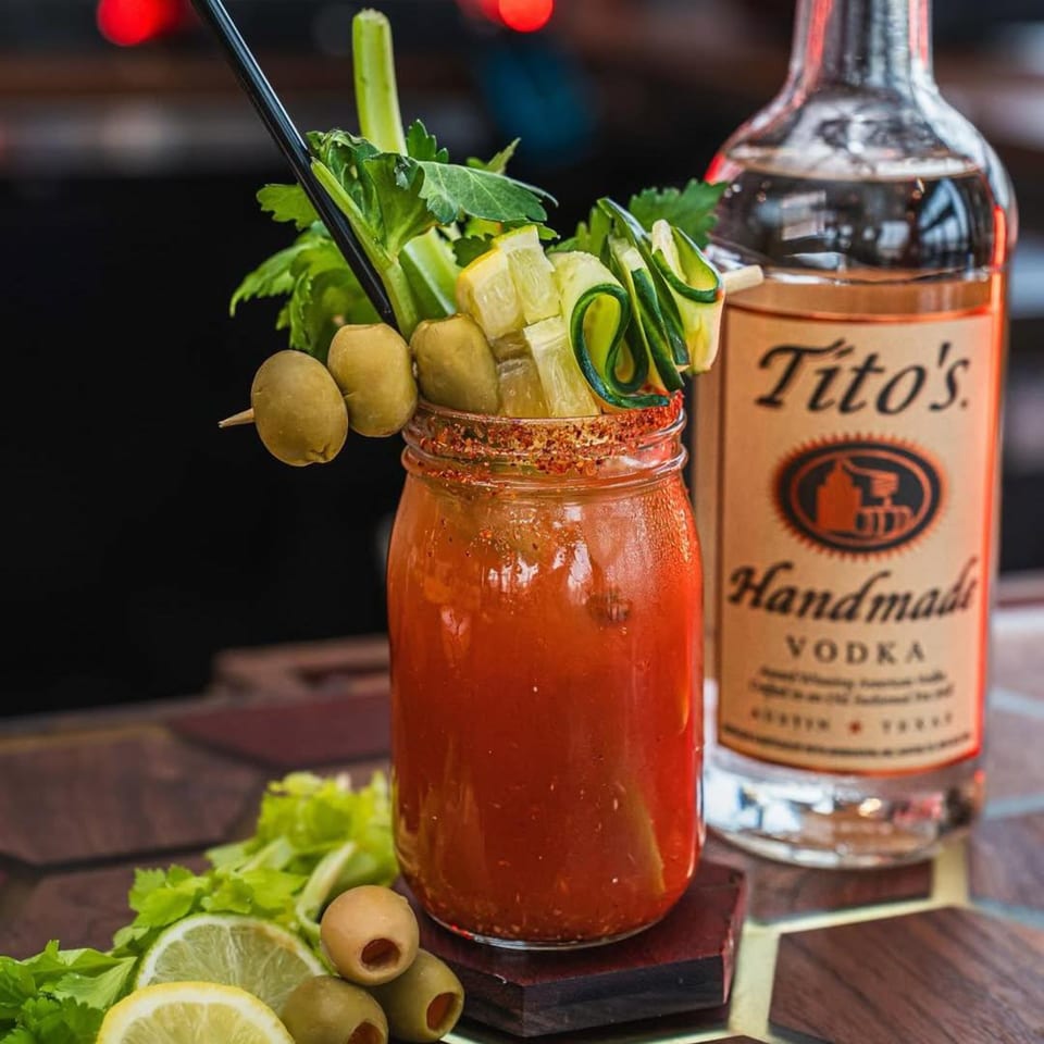 A Bloody Mary cocktail in a mason jar, garnished with olives, celery, cucumber, and lime, next to a bottle of Tito's Handmade Vodka.